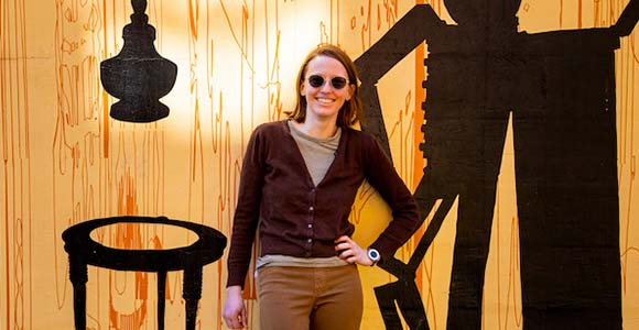 a woman standing in front of a yellow background with sunglasses, a brown cardigan, and light brown pants.